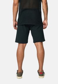 Shorts noirs à coupe décontractée, en tissu doux. Portés avec un haut en mesh noir et des baskets vert olive avec des accents rouges.