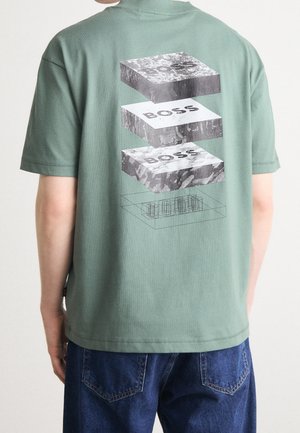 Osoba ubrana w zieloną koszulkę z krótkim rękawem z nadrukiem trójwymiarowych bloków ułożonych jedno na drugim z napisem "BOSS" na plecach, w połączeniu z niebieskimi dżinsami.