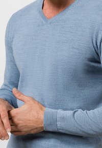 Jasnoniebieski sweter w serek z miękkiego materiału dzianinowego. Posiada ściągacze przy mankietach oraz gładką teksturę, a także dopasowane rękawy i korpus.
