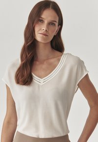 Blouse blanche à manches courtes avec un décolleté en V, avec deux lignes de couture décoratives le long du col. Fabriquée en tissu léger et soyeux.
