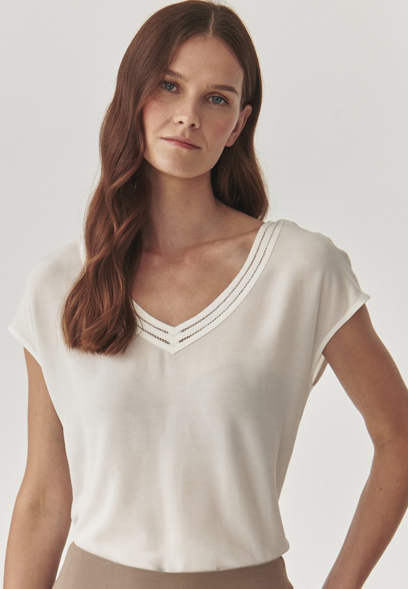 Blouse blanche à manches courtes avec un décolleté en V, avec deux lignes de couture décoratives le long du col. Fabriquée en tissu léger et soyeux.