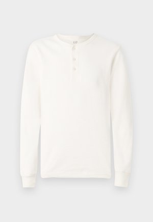 Bela majica s long sleeves in henley izrezom, s tremi gumbi in rebrastimi manžetami, prikazana na enostavni svetli podlagi.