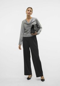 Blusa in satin grigio con scollatura a barchetta, abbinata a pantaloni neri a righe e tacchi a punta neri, mentre tiene una pochette nera.