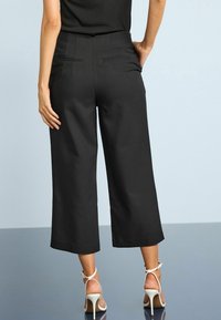 Pantalons larges noirs en tissu lisse, dotés de poches arrière et d'une coupe ajustée, associés à des sandales à talons hauts de couleur claire.