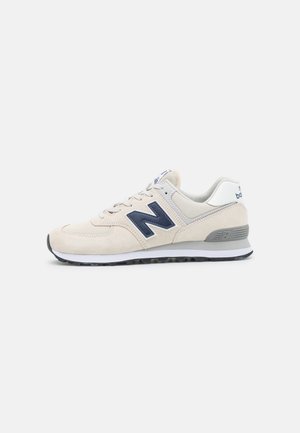 Hellbeiger New Balance Sneaker mit navy Logo, weißer und grauer Sohle und Schnürverschluss auf weißem Hintergrund.