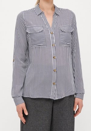 Femme portant une chemise à boutons à rayures verticales bleu marine et blanches à manches longues avec deux poches poitrine et un pantalon gris foncé.