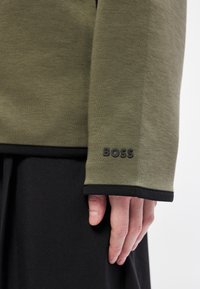 Dlhý olivovo zelený top s čiernymi manžetami a lemom, s vystúpeným logom "BOSS" na manžete. Mäkká textúra, neformálny dizajn.