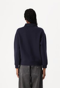 Marineblauwe sweatshirt met een kraag, geribde manchetten en een relaxte pasvorm; gecombineerd met grijze hoge taille broek met achterzakken.
