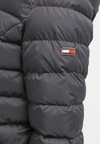 Černá nafukovací bunda s prošívaným designem, která má na rukávu malou látkovou nášivku v červené, bílé a modré barvě.