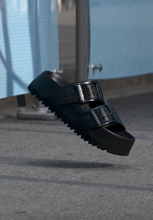 Sandalo nero con due fibbie regolabili, suola spessa e fondo scanalato, sospeso sopra un marciapiede grigio in un ambiente esterno.