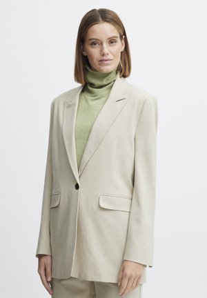 Beige Blazer mit strukturierten Revers, Ein-Knopf-Verschluss und zwei Fronttaschen. Getragen über einem hellgrünen Rollkragenpullover. Glatte Stofftextur.