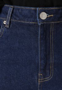 Μπλε τζιν παντελόνι σε σκούρο χρώμα, με ψηλή μέση, ασημένιο κουμπί και κίτρινη ραφή κατά μήκος των ραφών και των τσεπών.