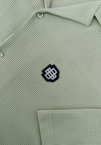 Lichtgroene geprofileerde polo met een drieknops sluiting, een patchzak en een donkere geborduurde logo op de borst.