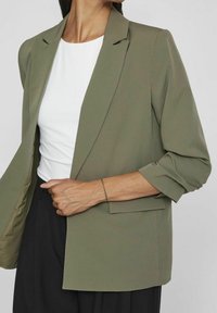 Veste olive verte avec manches retroussées, col cranté et fermeture à un bouton. Portée sur un haut blanc, avec un design minimaliste.