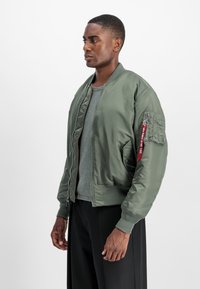 Jonge man in een olijfgroene bomberjack met een rode "Remove Before Flight" label op de mouwzak, grijs shirt en zwarte broek, staand in zijaanzicht.