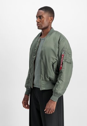 Young man wearing an olive green bomber jacket with a red "Remove Before Flight" tag on sleeve pocket, gray shirt, and black pants, standing sideways.