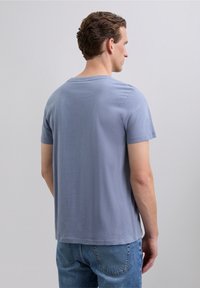 Mann, der sich abwendet und ein hellblaues kurzärmeliges T-Shirt sowie blaue Jeans trägt, vor einem einfarbigen grauen Hintergrund.