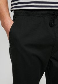 Czarne casualowe spodnie z elastycznym paskiem i sznurkiem, z kieszeniami bocznymi i gładką, matową fakturą. Widoczna jest paskowana koszula powyżej.