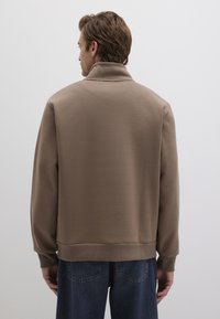 Brown sweatshirt with a high collar and ribbed cuffs, featuring a soft texture and relaxed fit. Worn over dark blue jeans.
