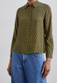 Camicia a botón in tessuto nero con un motivo ripetuto di "M" gialla. Maniche lunghe, colletto e orlo dritto. Indossata con jeans blu.