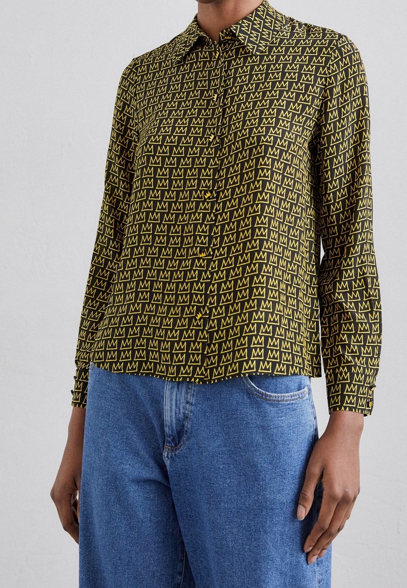 Camicia a botón in tessuto nero con un motivo ripetuto di "M" gialla. Maniche lunghe, colletto e orlo dritto. Indossata con jeans blu.