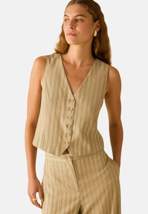 Kvinde iført beige ærmeløs vest med nålestriber og matchende højhælspantaloner, med guldbelagte øreringe og ring, stående med hånden i lommen.