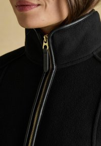 Detailný záber na osobu oblečenú v čiernej fleecovej bunde so zlatým zipsom a golierom z kože, zipsovanou až po bradu.