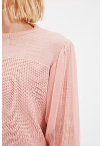 Ljusrosa tröja med stickad kropp och genomskinliga puffärmar. Tyget har en strukturerad yta med ett horisontellt randmönster över bröstet.