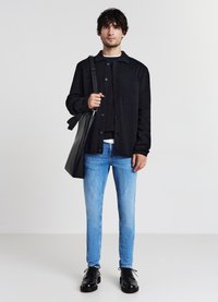 Schwarze Knopfjacket über einem schwarzen Pullover, hellblaue Jeans und schwarze Schnürschuhe. Eine schwarze Tote-Bag wird in der linken Hand gehalten.