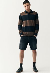 Gestreiftes langärmliges Polo in Braun und Marineblau, kombiniert mit dunklen Denim-Shorts. Das Model trägt marineblaue Socken und schwarze Schuhe mit braunen Sohlen.