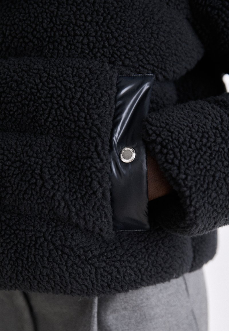 Giacca in pile nera con una texture morbida e un accento nero lucido ai polsini, fissata con un bottoncino metallico.