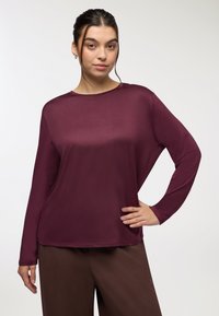 Camicia bordeaux a maniche lunghe con una vestibilità comoda, texture liscia, scollatura rotonda e una caduta elegante. Abbinata a pantaloni ampi marroni.