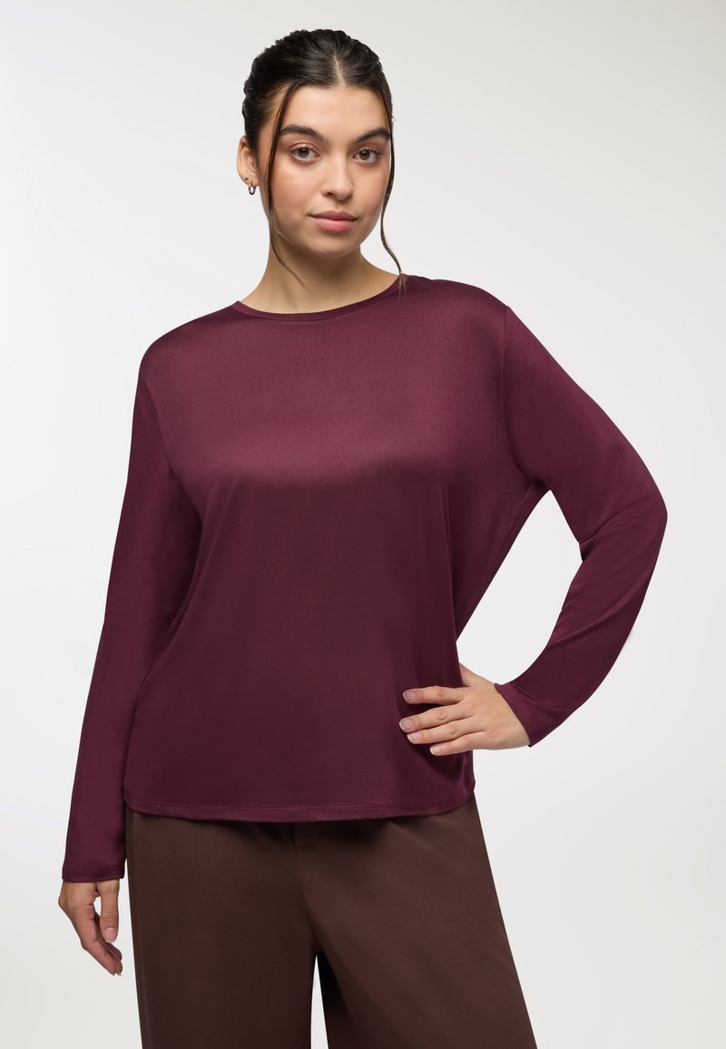 Camicia bordeaux a maniche lunghe con una vestibilità comoda, texture liscia, scollatura rotonda e una caduta elegante. Abbinata a pantaloni ampi marroni.