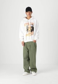 Camisola branca com uma graphic impressa de um homem e o texto "Thug Life California Love", combinada com calças largas em verde-azeitona e ténis brancos.