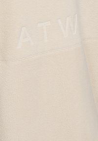 Krämfärgad fleece-material med en slät textur, med en söm som är sydd och präglad "ATW"-bokstäver på ytan.
