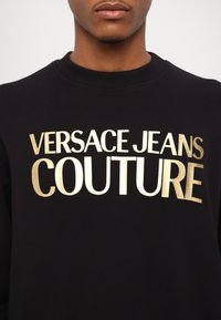 Felpa nera con scollo rotondo e il bold "VERSACE JEANS COUTURE" in lettere dorate metallizzate stampato sul davanti.