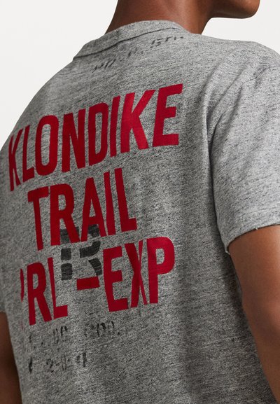 Harmaa t-paita, jonka selässä on rohkeaa punaista tekstiä "KLONDIKE TRAIL PRL-EXP". Pehmeä kangas, pyöreä kaula-aukko ja lyhyet hihat.