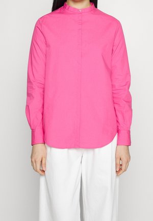 Personne portant une chemise rose vif à manches longues avec col mandarin et un pantalon blanc ample.