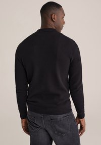 Črn pleten pulover z teksturirano površino, z dolgimi rokavi, okroglo izrezom in rebrastimi manšetami ter spodnjim robom. Nošen z temnimi kavbojkami.