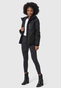 Black insulated jacket with a zippered front, detachable hood, and side pockets. Worn over a white top and paired with dark jeans and ankle boots.