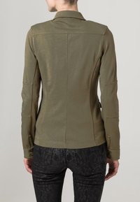 Olive green fitted jacket with a collar, stitched detailing, and button accents. Paired with patterned black pants for contrast.