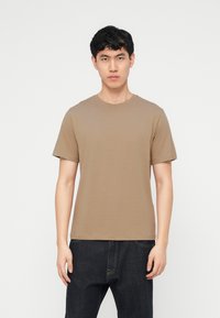 Νεαρός άνδρας που φοράει ένα απλό μπεζ κοντομάνικο μπλουζάκι και σκούρο τζιν, στέκεται μπροστά σε ένα λευκό φόντο.