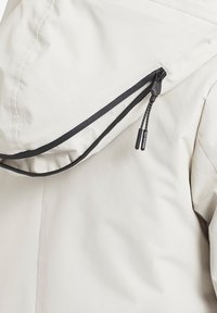 Vaaleanbeige takki, jossa on huppu mustilla yksityiskohdilla, ulkopuolella vetoketju vetonauhalla ja sileä, vedenpitävä kangas.