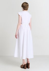 Donna che indossa un vestito midi bianco senza maniche con dettagli cut-out sulla schiena e sandali con tacco a pianta piatta marroni, in piedi davanti a uno sfondo neutro.