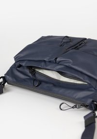 Marineblaue gepolsterte Tasche mit mehreren schwarzen Reißverschlussanhängern, ein Reißverschluss teilweise geöffnet, der das helle Innenfutter zeigt, und verstellbarem Gurt.