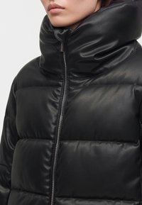 Gros plan d'une personne portant une veste noire matelassée avec un col montant et une fermeture éclair à l'avant, lèvres visibles, visage partiellement montré.