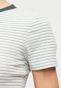 Gros plan sur une personne portant un t-shirt blanc à manches courtes côtelé avec de fines rayures horizontales noires et un col foncé.