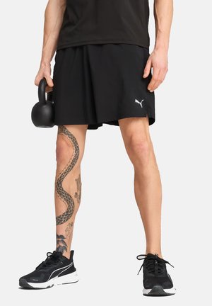 Schwarze Sportshorts mit einem weißen Logo, schwarze Sportschuhe und eine Kettlebell; das Bein zeigt ein detailliertes Schlangentattoo.
