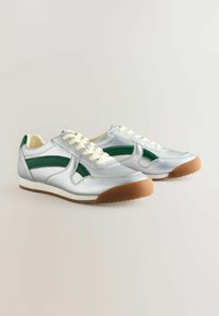 Baskets de sport argentées avec des accents verts, présentant une tige texturée, des lacets contrastants et une semelle en caoutchouc avec un profil marron.