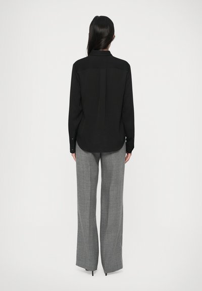 Frau mit langen dunklen Haaren, die ein schwarzes Langarmshirt und graue weite Hosen trägt, steht vor einem weißen Hintergrund.